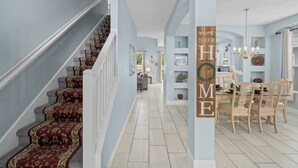Un vaste hall d’entrée agrémenté d’un magnifique escalier, un coin repas avec des chaises en bois, et une pancarte accueillant les invités dans la maison.