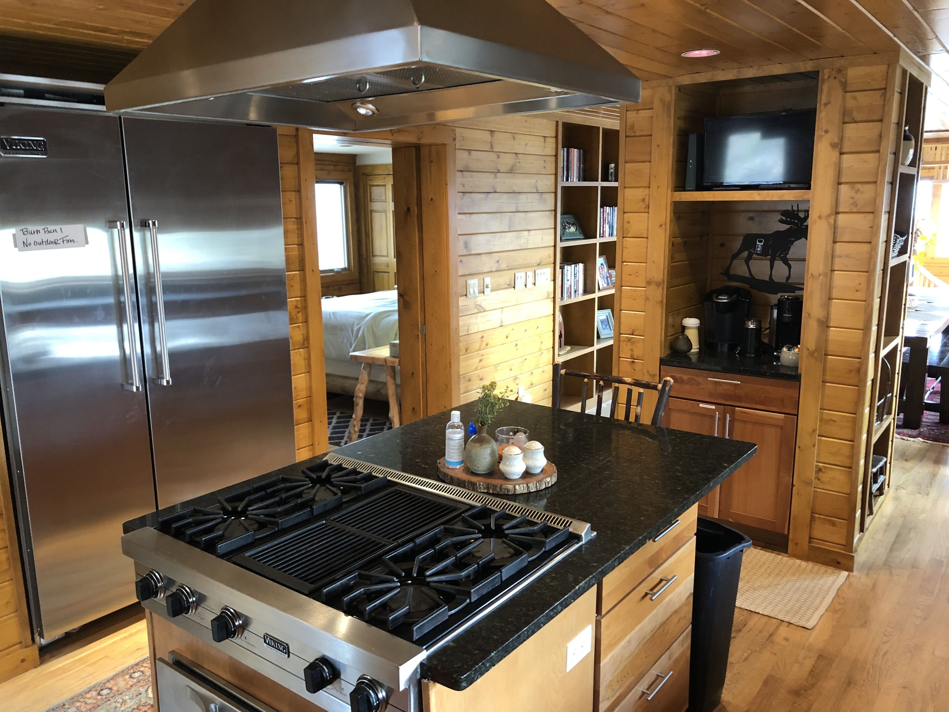 Una cocina compacta con un refrigerador de acero inoxidable, estufa a gas, armarios de madera y una estantería empotrada.