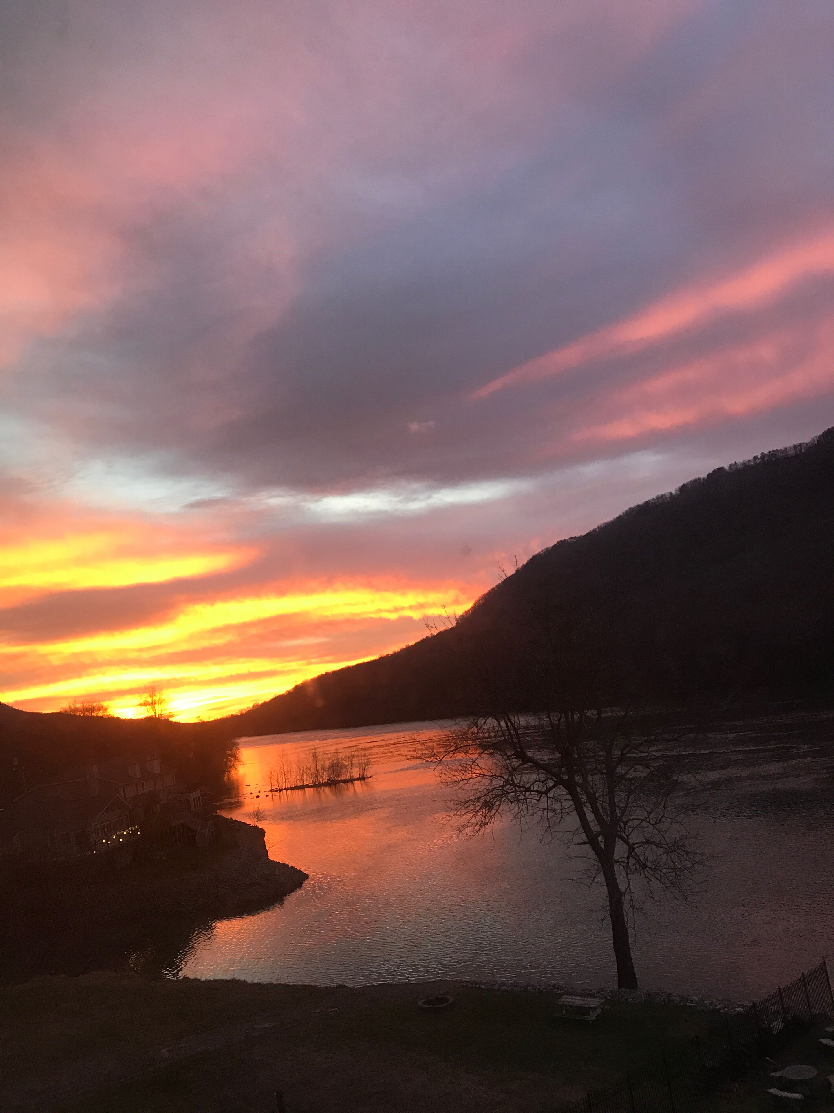 A sunset over a calm lake with a silhouette of a hill and a house on the left.