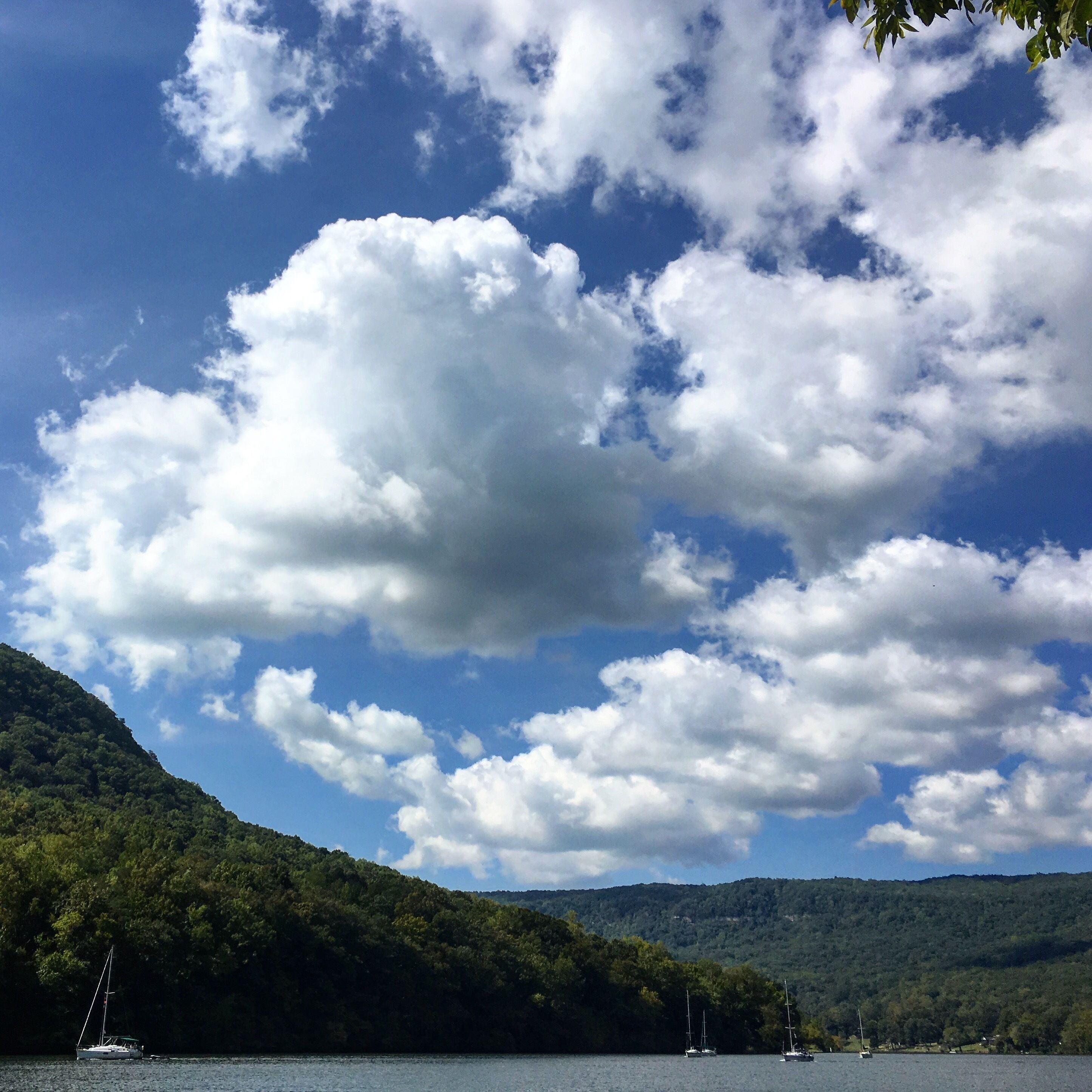 A lake with sailboats, surrounded by forested hills under a partly cloudy sky.