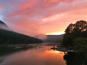 A calm river with boats docked on the right, surrounded by forested hills and a sunset sky.