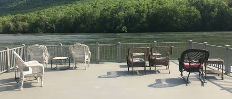 A deck with wicker chairs and a table overlooking a lake, surrounded by a railing and a forested hillside.