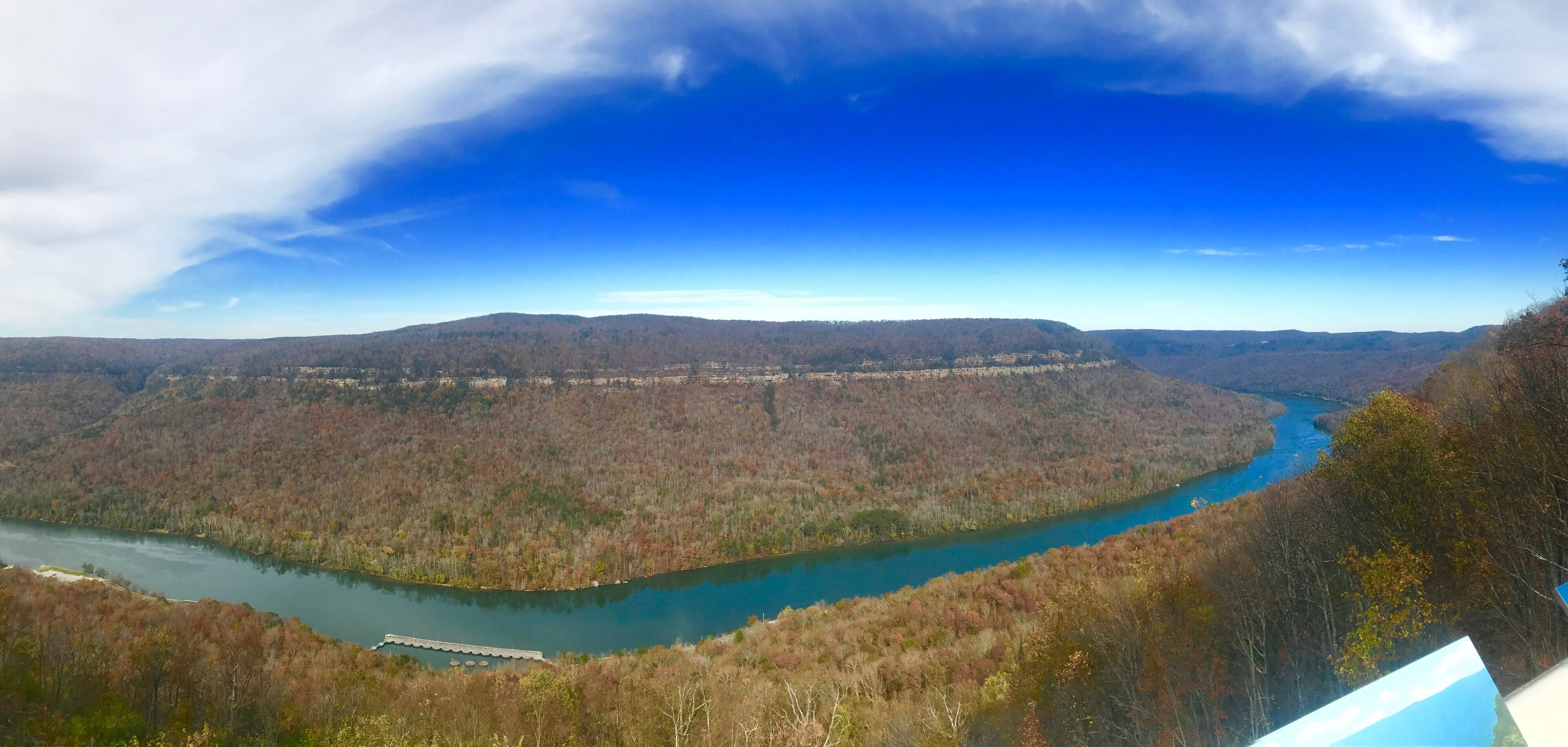 Une vue panoramique d’une rivière serpentant à travers une vallée boisée, avec un pont et des collines lointaines.