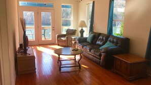 A spacious living room with hardwood floors, a leather sofa, a patterned armchair, a small table, and a cabinet. There are two windows with curtains, a ceiling fan, and a door leading to an outdoor area. - Waterfront Paradise- 10 minutes from downtown Chattanooga (Chattanooga)