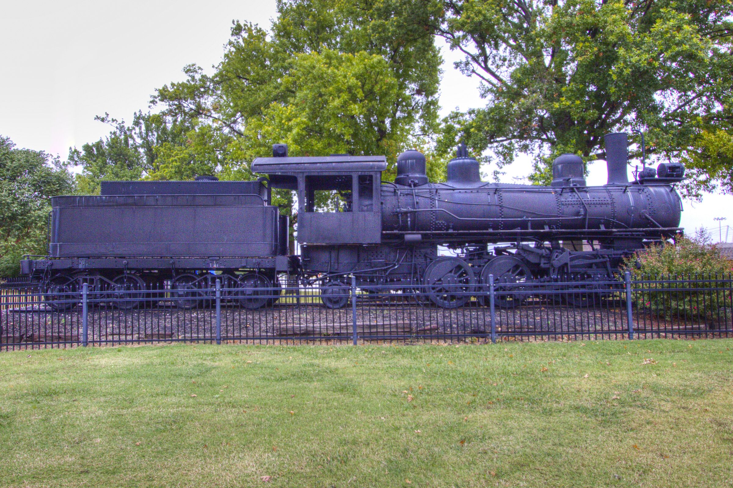 Une locomotive à vapeur immobilisée, avec un wagon à plateau, entourée d’une clôture et d’arbres.