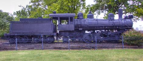 Une locomotive à vapeur immobilisée, avec un wagon à plateau, entourée d’une clôture et d’arbres.