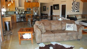 A spacious living area with a brown leather sofa, wooden coffee table, and a cowhide rug. A kitchen with wooden cabinets and stainless steel appliances is visible in the background. - Lake front home private dock (Paso Robles)
