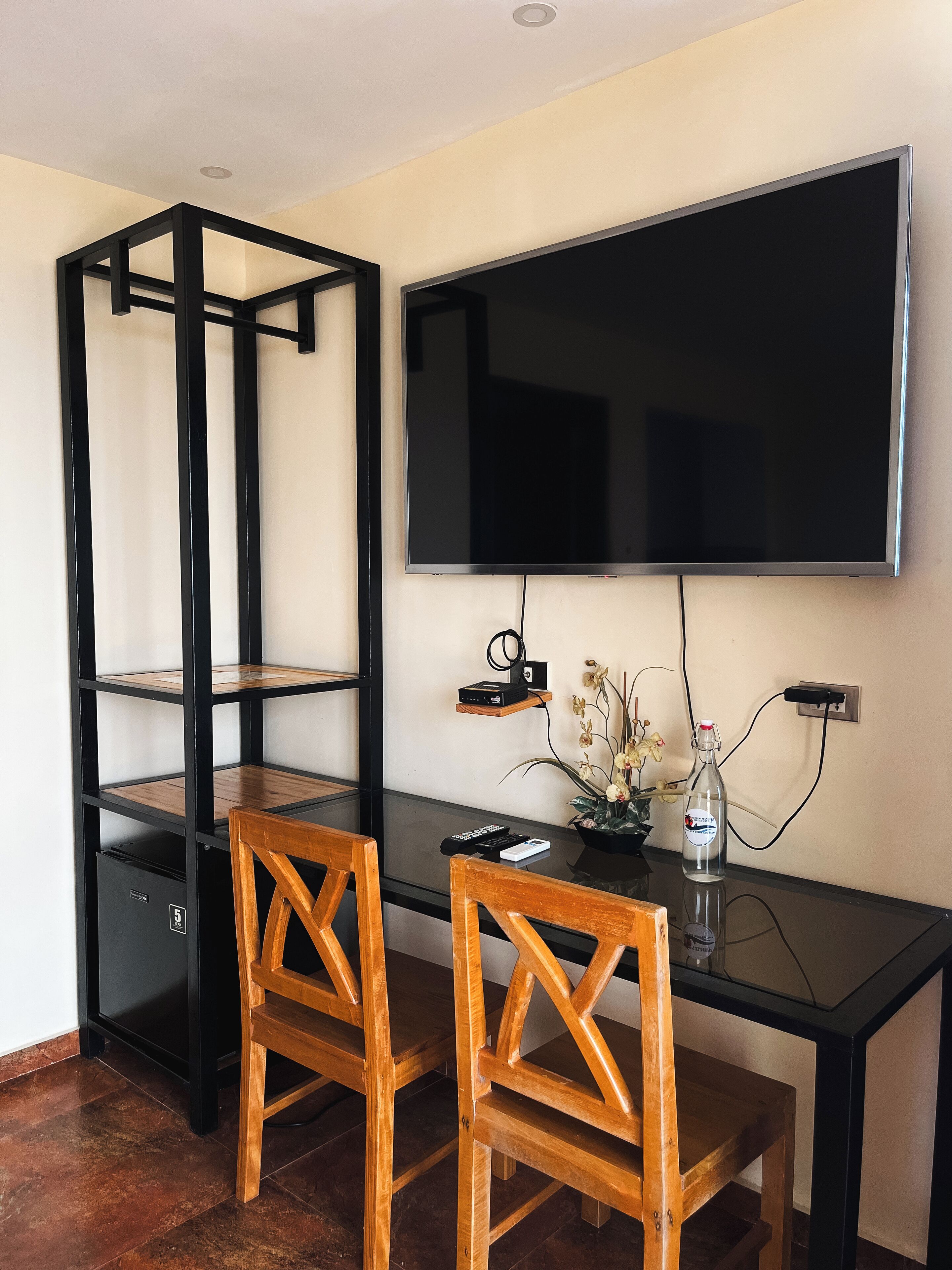 Una habitación moderna con un escritorio de vidrio, dos sillas de madera, un televisor negro en la pared, un estante pequeño con un altavoz y un florero con flores.
