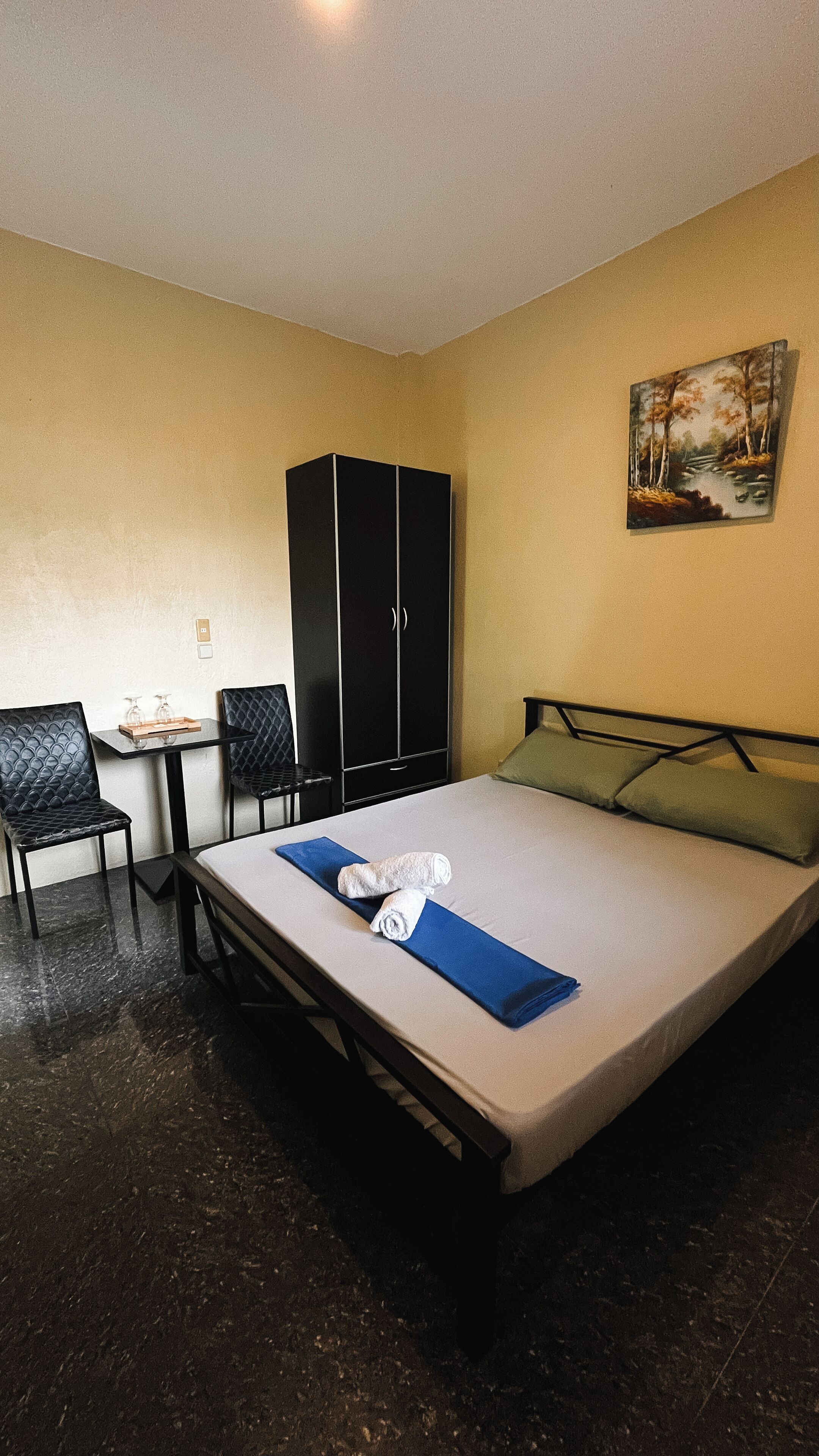 Una cama individual con una toalla azul y un almohadón blanco, un armario negro, una mesita con dos vasos y un cuadro en la pared.