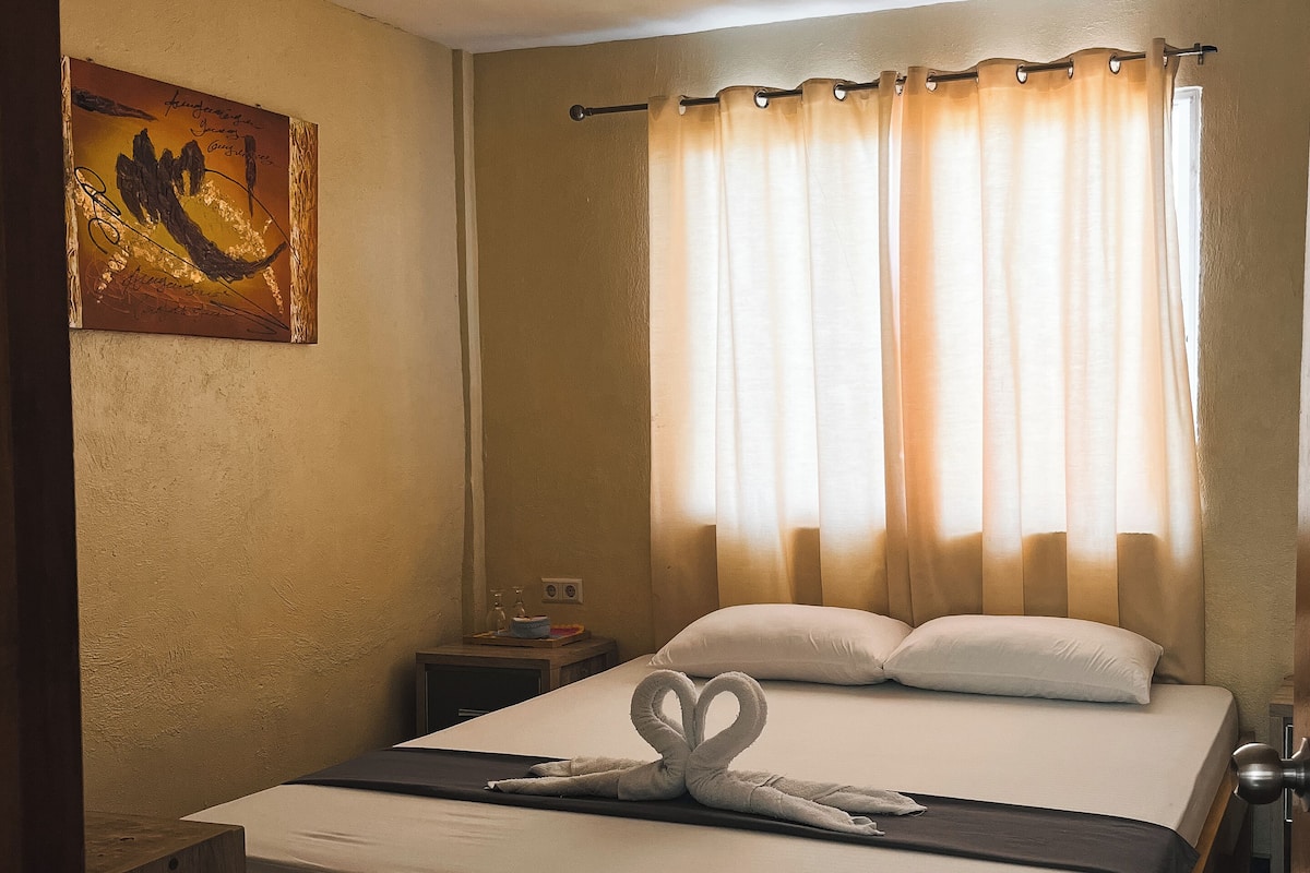 Quarto de hotel com uma cama de madeira, uma mesa de cabeceira com luminária, um quadro na parede e cortinas translúcidas.