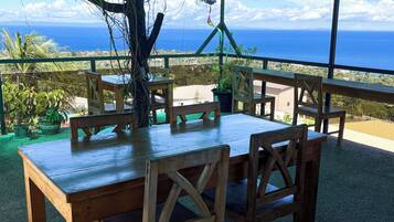 An outdoor dining area with wooden tables and chairs, a tree, and a view of the ocean.