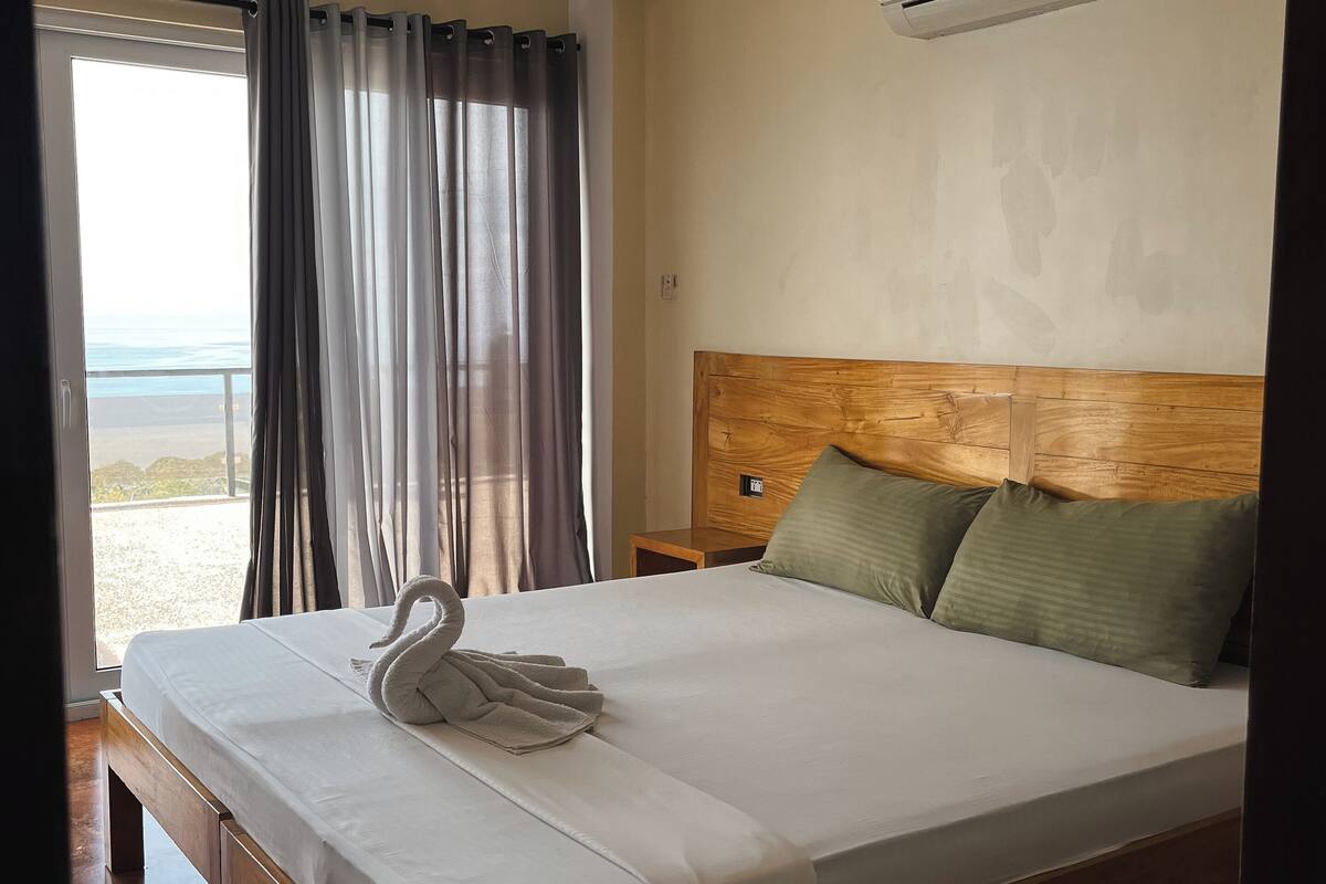 Quarto de hotel com uma cama grande, cabeceira de madeira, vista para o mar pela janela e um aparelho de ar condicionado na parede.