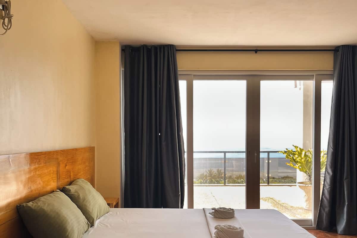 Quarto com uma cama grande, cabeceira de madeira e vista para o oceano através de uma porta de correr de vidro.