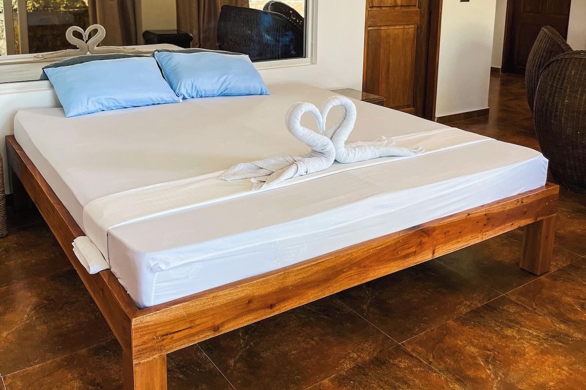 Uma cama bem arrumada com uma toalha em formato de coração, uma janela grande com cortinas e uma porta de madeira.