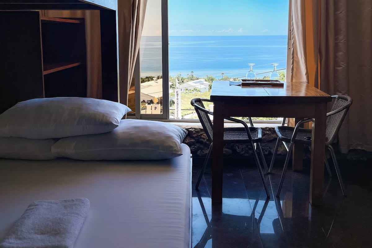 Um quarto com uma cama de solteiro, uma mesa com cadeiras e vista para o mar.