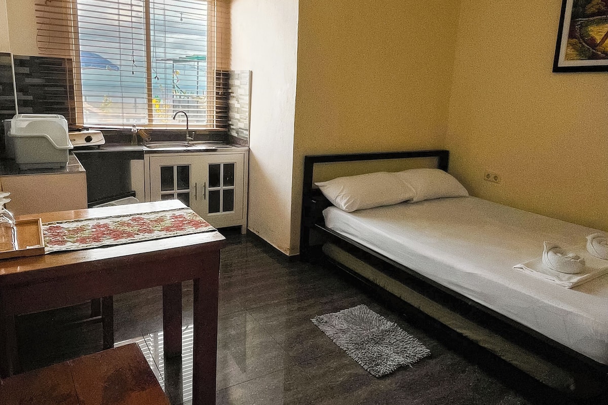 Um quarto pequeno e pouco mobiliado, com uma cama, uma mesa, uma janela com persianas e uma cozinha compacta.