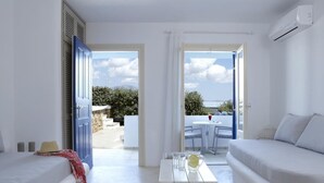 A bright, airy room with a white sofa, a small table with glasses and a pitcher, and a view of the outdoors through an open door. - Hotel Mediterranean (Naxos)