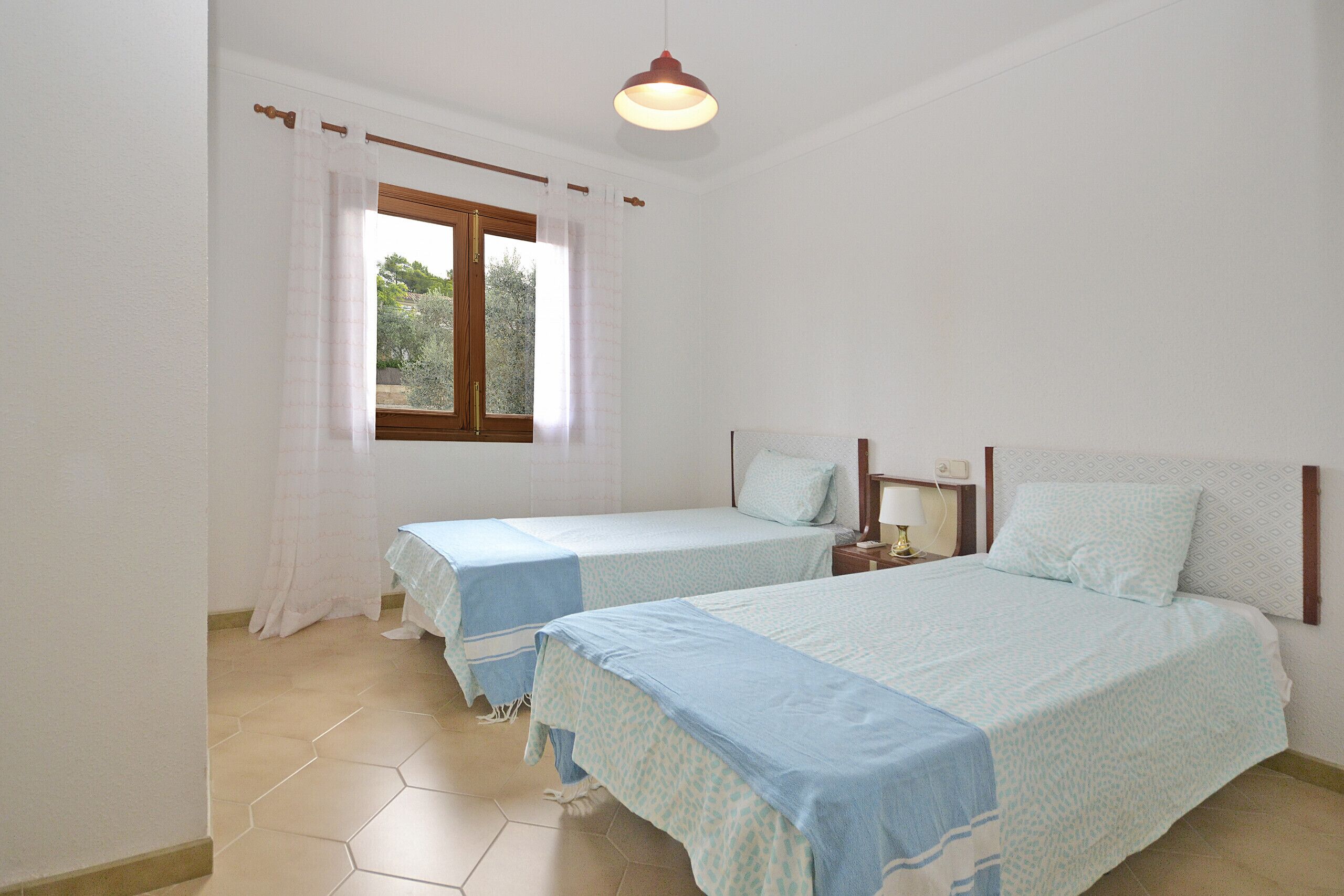 A bedroom with two beds, a window with curtains, a hanging light fixture, and a tiled floor.