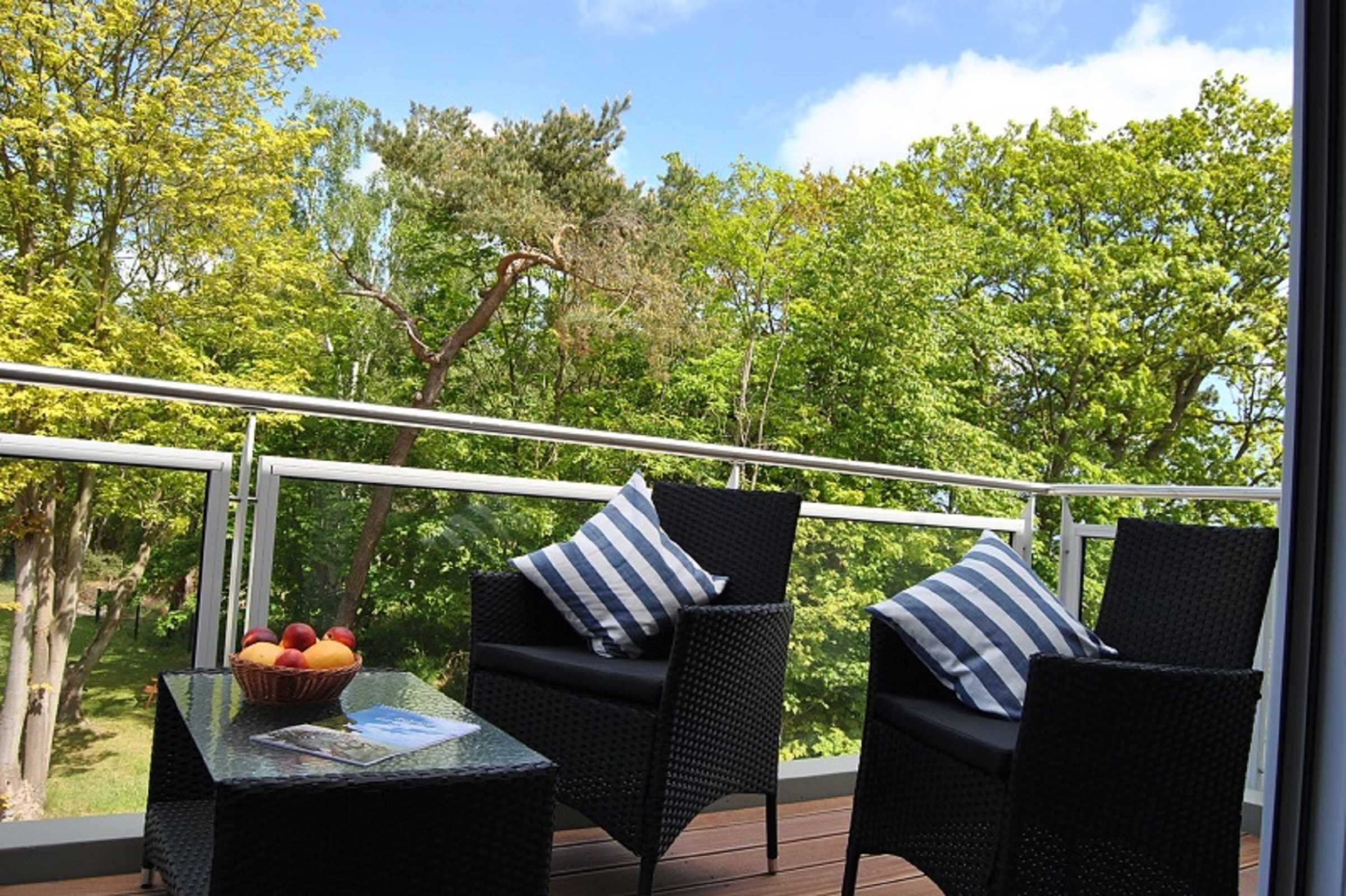 Een balkon met rieten meubels, een tafel met een fruitmand, en uitzicht op groene bomen.