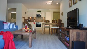 Ein modernes Wohnzimmer mit einer Couch, einem Couchtisch mit Früchten, Holzstühlen, einem Fernseher und einer Küche im Hintergrund.
