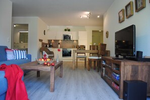 Una sala de estar moderna con un sofá, una mesa de centro con fruta, sillas de madera, un televisor y una cocina al fondo.