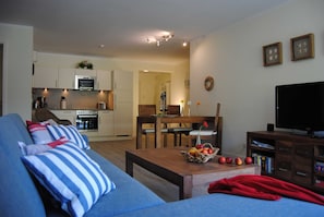 Uma sala de estar com sofá, mesa de centro com frutas, televisão e cozinha ao fundo.