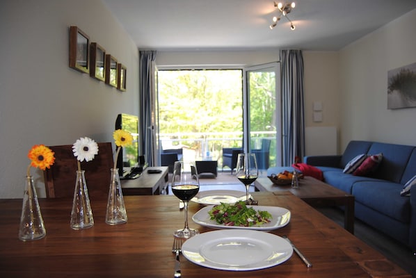 Uma mesa de jantar com pratos, copos de vinho e vasos com flores. Há um sofá azul e uma área de refeições com vista para o exterior.