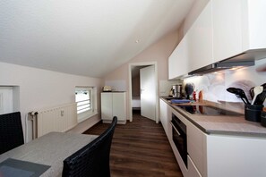 Une cuisine de taille réduite avec des meubles blancs, un plancher en bois, un coin repas comprenant une table et des chaises, et une vue sur une chambre à coucher par une porte ouverte.