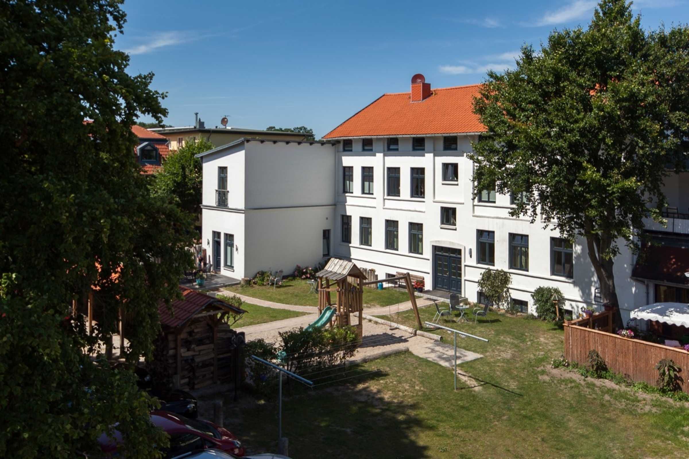 A courtyard with a playground, a white building with a red roof, and a wooden playhouse.