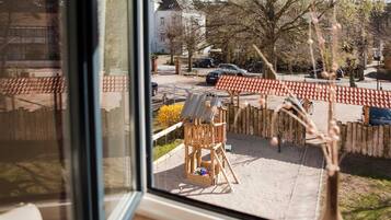 Blick aus einem Fenster auf einen Spielplatz mit einer Holzkonstruktion und Sand.