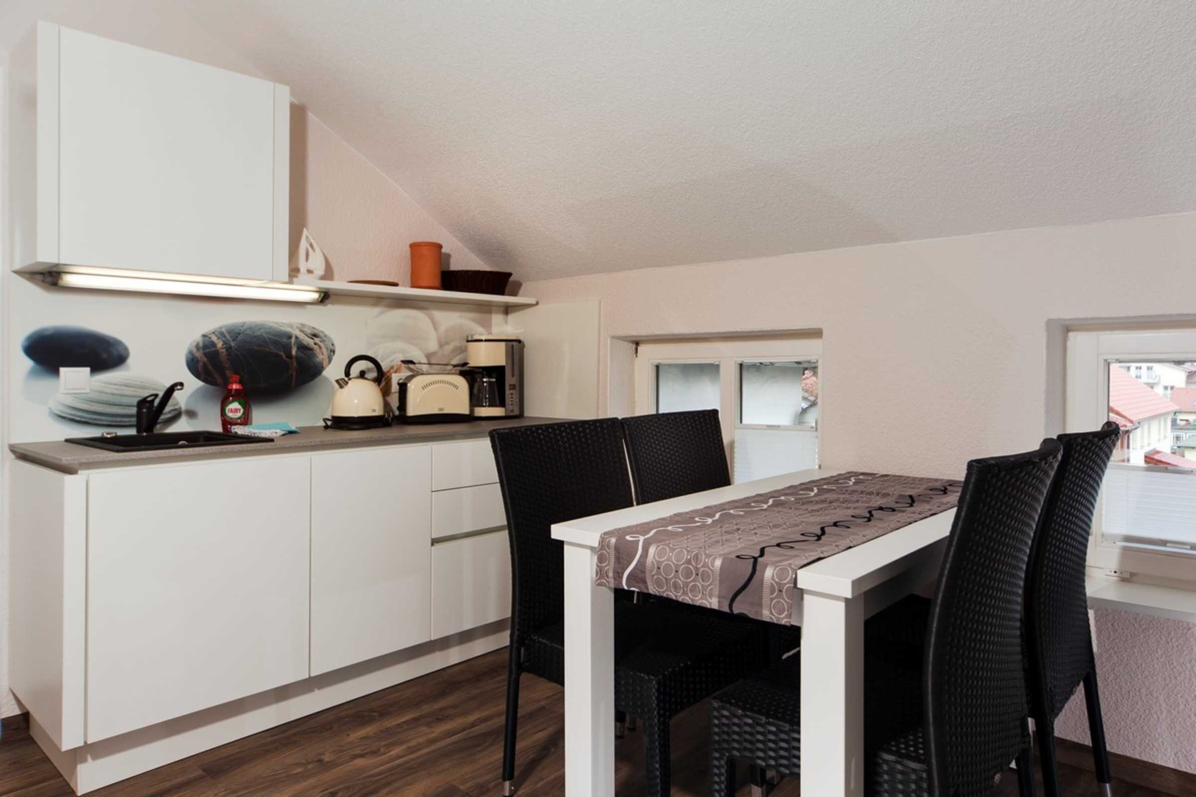 Cocina compacta con armarios blancos, cafetera y comedor con mesa y sillas.