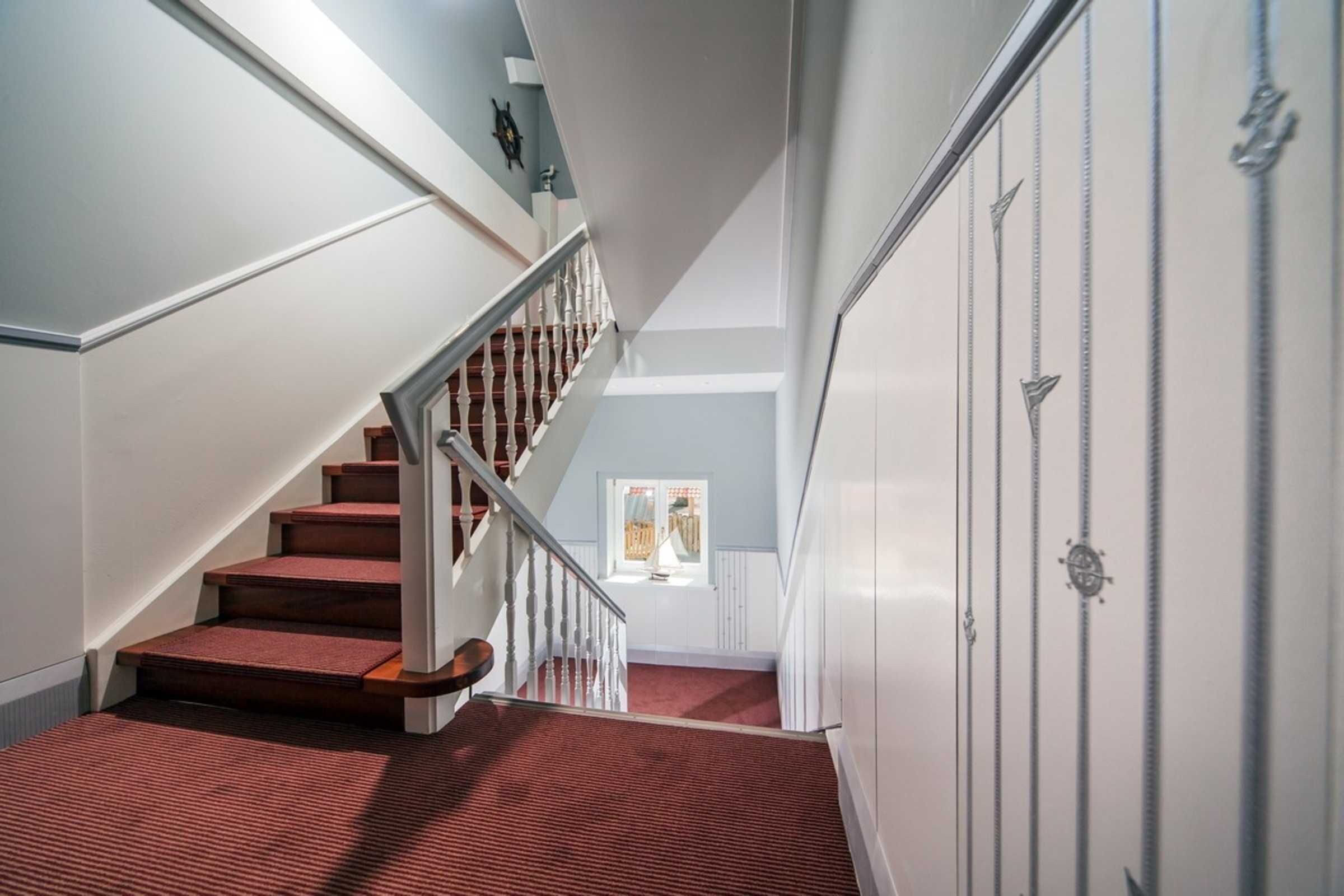 Un escalier avec des marches en bois et une moquette rouge, menant à une pièce dotée d’une fenêtre et d’une armoire blanche.