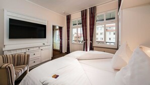 A hotel room with a large flat-screen TV mounted on the wall, a bed with white linens, a striped armchair, and a view of buildings through the windows.
