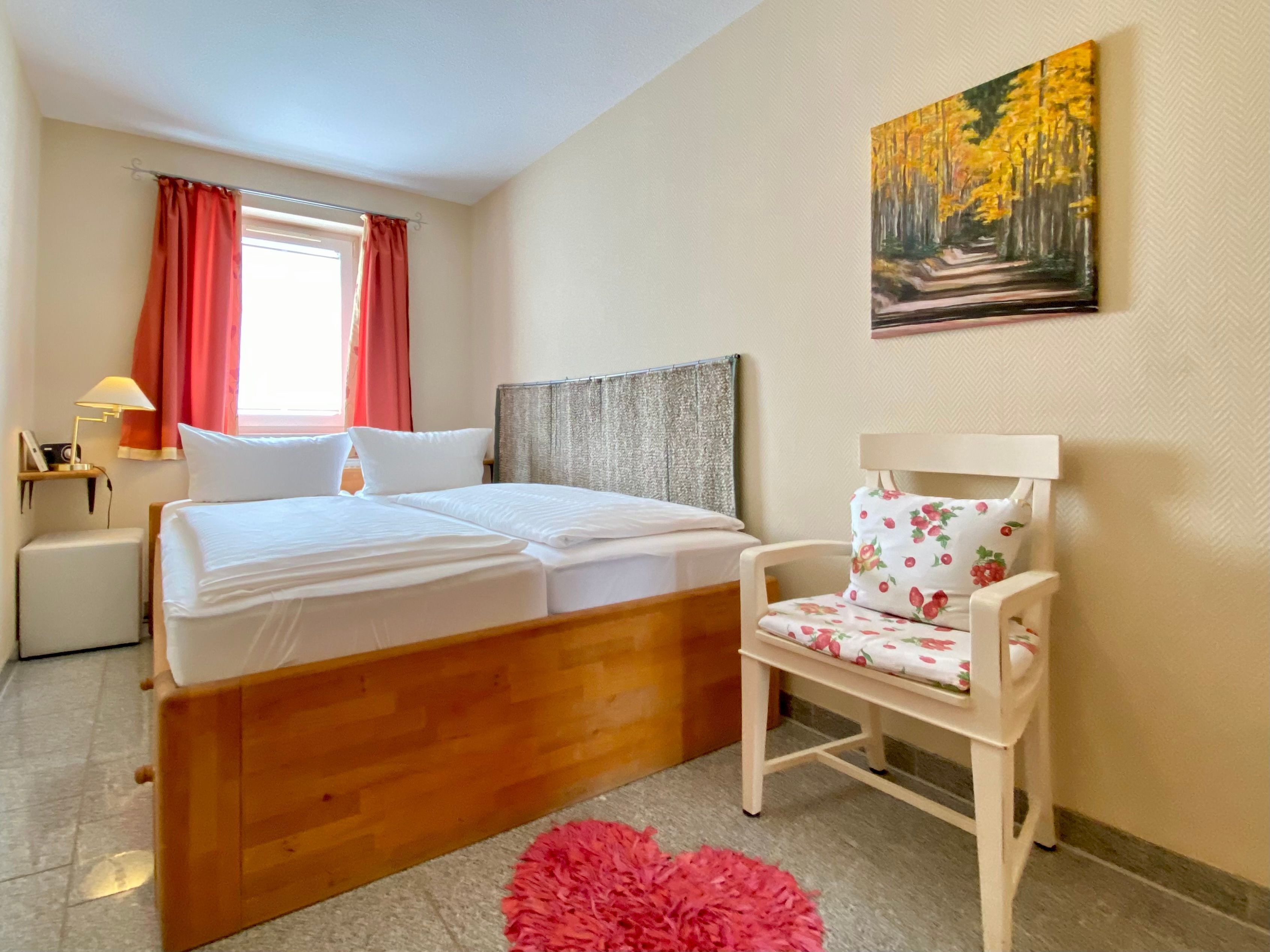 A hotel room with a wooden bed, a chair with floral upholstery, a painting of a forest, and a window with red curtains.