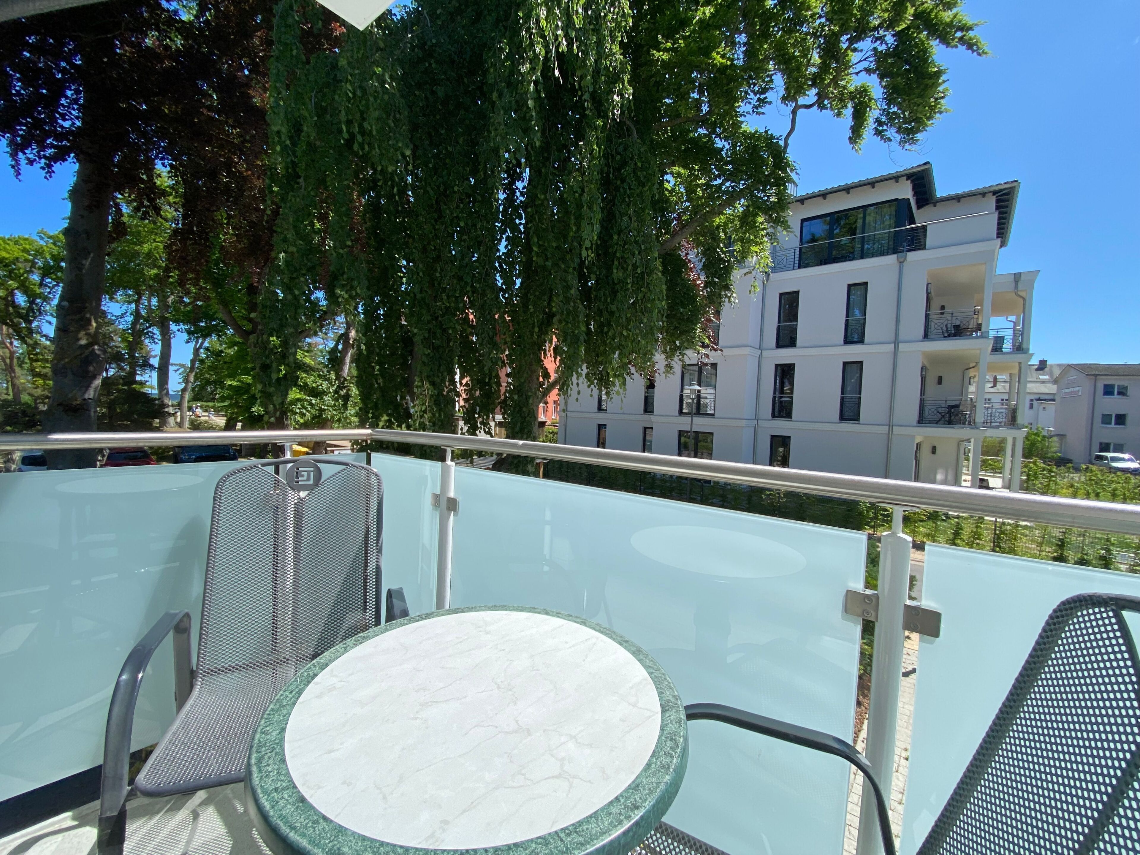 Varanda com mesa e cadeiras de vidro, vista para um edifício moderno e árvores.
