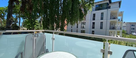 A balcony with a glass table and chairs, overlooking a modern building and trees.