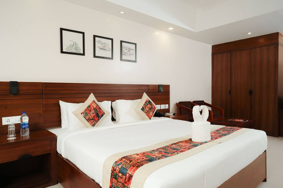 A hotel room with a wooden headboard, white bedding, a decorative pillow, a water bottle, a bedside table, and a wooden wardrobe.