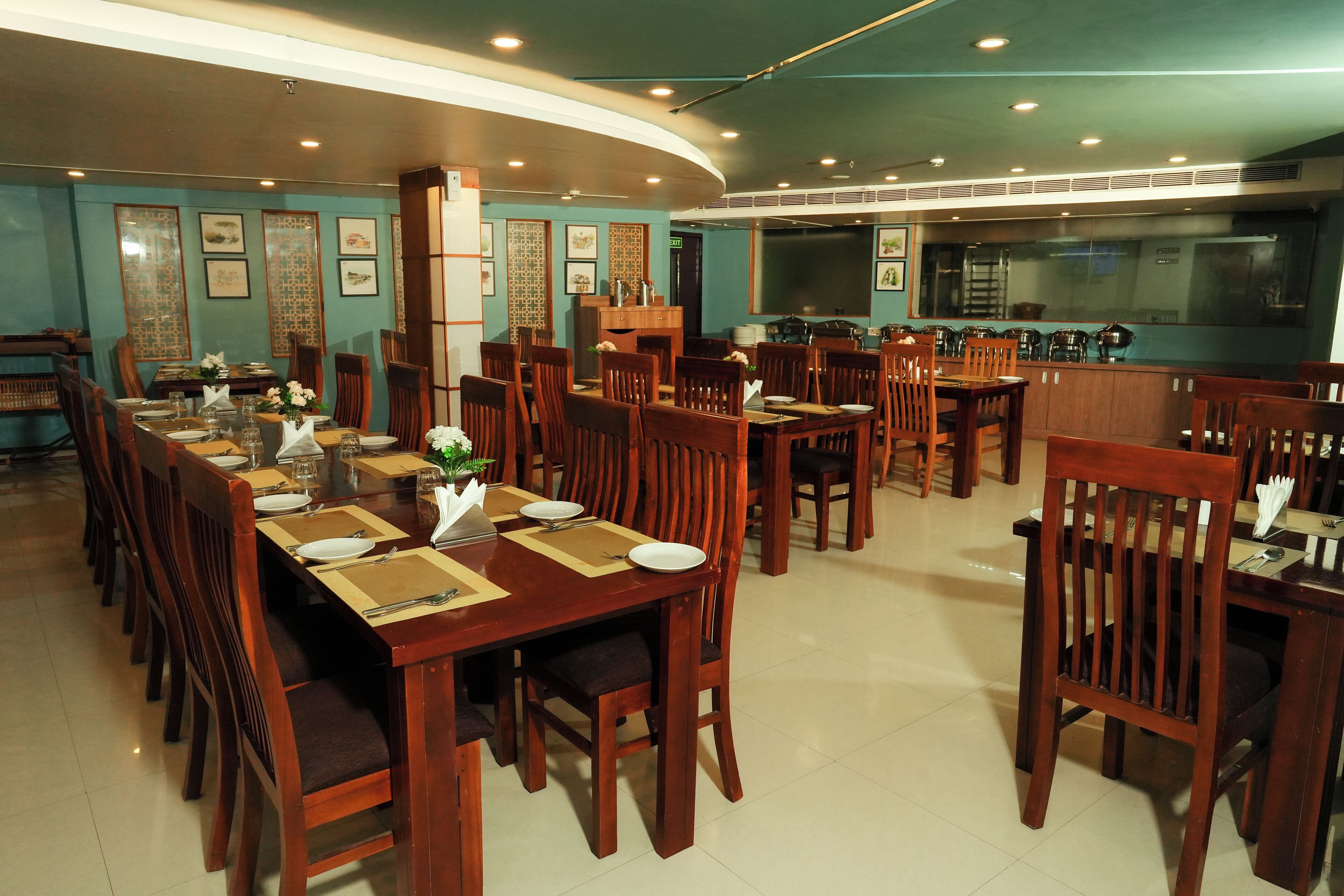 A restaurant with wooden tables and chairs, a kitchen area, and framed artwork on the walls.