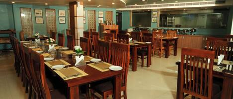 A restaurant with wooden tables and chairs, a kitchen area, and framed artwork on the walls.