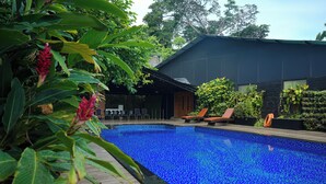 A swimming pool with a wooden deck, surrounded by lush greenery and a modern building with a dark facade. - Dfort Hotel Kollam (Kollam)