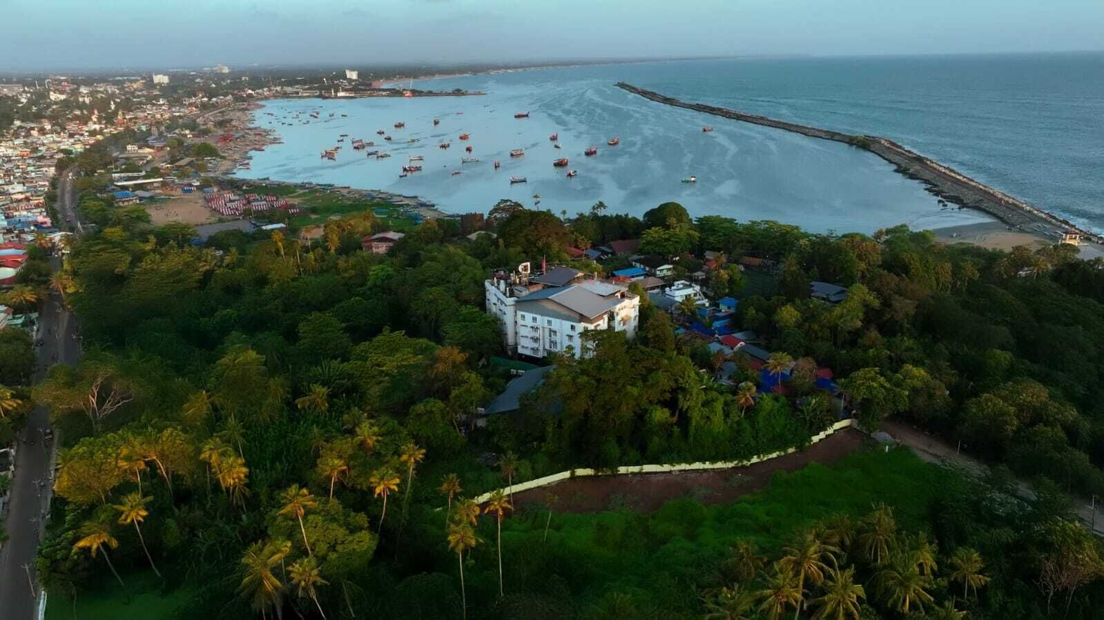 Luchtbeeld van een kustgebied met een opvallend wit gebouw, omringd door weelderige begroeiing en een strand vol schepen.