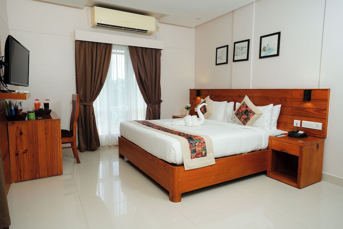 A hotel room with a wooden bed, a bedside table with a phone, a TV on a stand, and a desk with a computer and other items.
