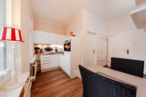 A compact kitchen with white cabinets, a built-in oven, and a dining area with a black chair. - Holiday home Wieck - Haus Grahl GM 69912 (Graal-Müritz)
