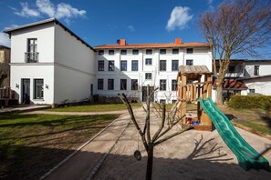 A playground with a slide and climbing structure, surrounded by a building with multiple windows and a red roof. - Holiday home Wieck - Haus Grahl GM 69912 (Graal-Müritz)