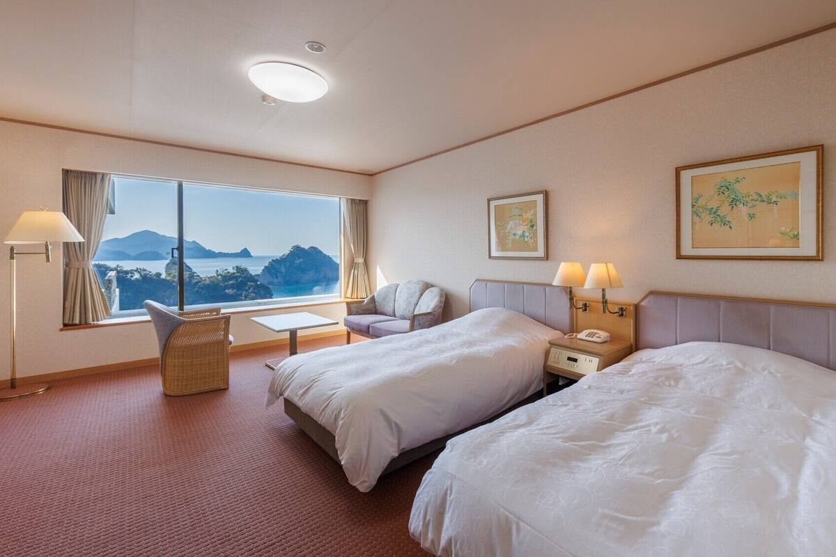 A hotel room with two beds, a desk, a chair, a lamp, and a window with a view.