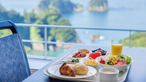 Bandeja de desayuno con cruasanes, ensalada, maíz y un vaso de jugo de naranja, sobre un balcón con vistas a un paisaje costero.