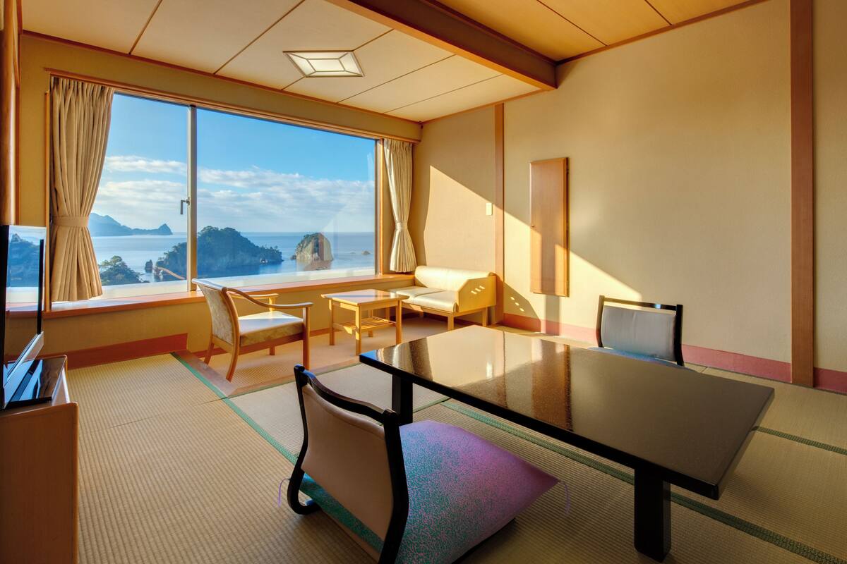 Une pièce dotée d’un sol recouvert de tatamis, d’une table basse, d’un canapé et d’une grande fenêtre offrant une vue sur un paysage côtier.