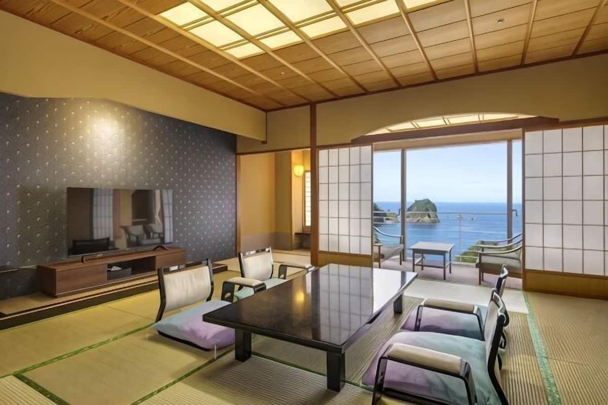 Um quarto japonês tradicional com tatames, uma mesa baixa, cadeiras, televisão e vista para o mar.