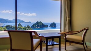 Habitación con un amplio ventanal que ofrece vistas a un paisaje costero con montañas y una masa de agua. Hay dos sillas de madera y una mesita en la habitación.