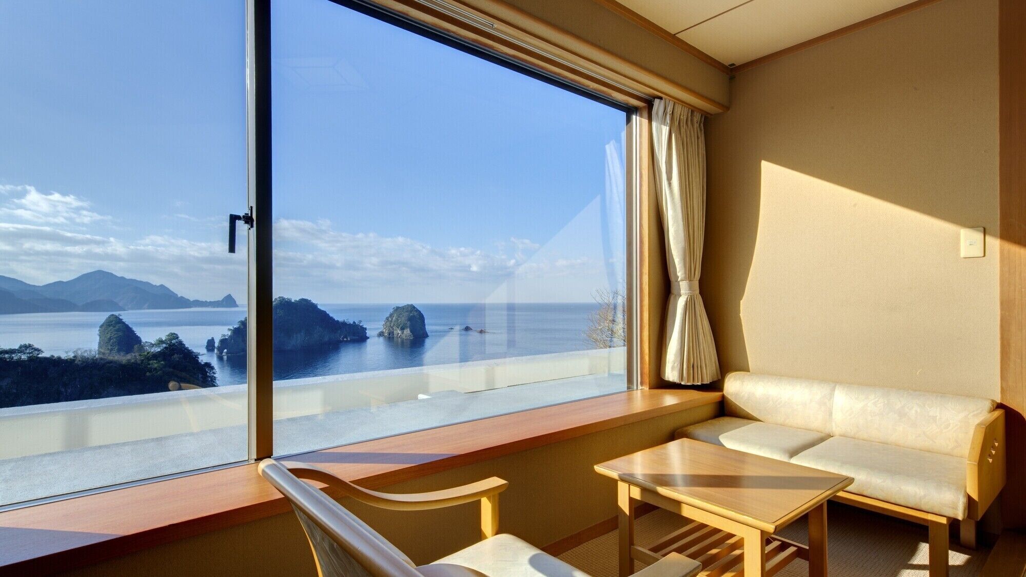 Een hotelkamer met een groot raam dat uitzicht biedt op zee en eilanden. Er is een bank en een klein tafeltje.